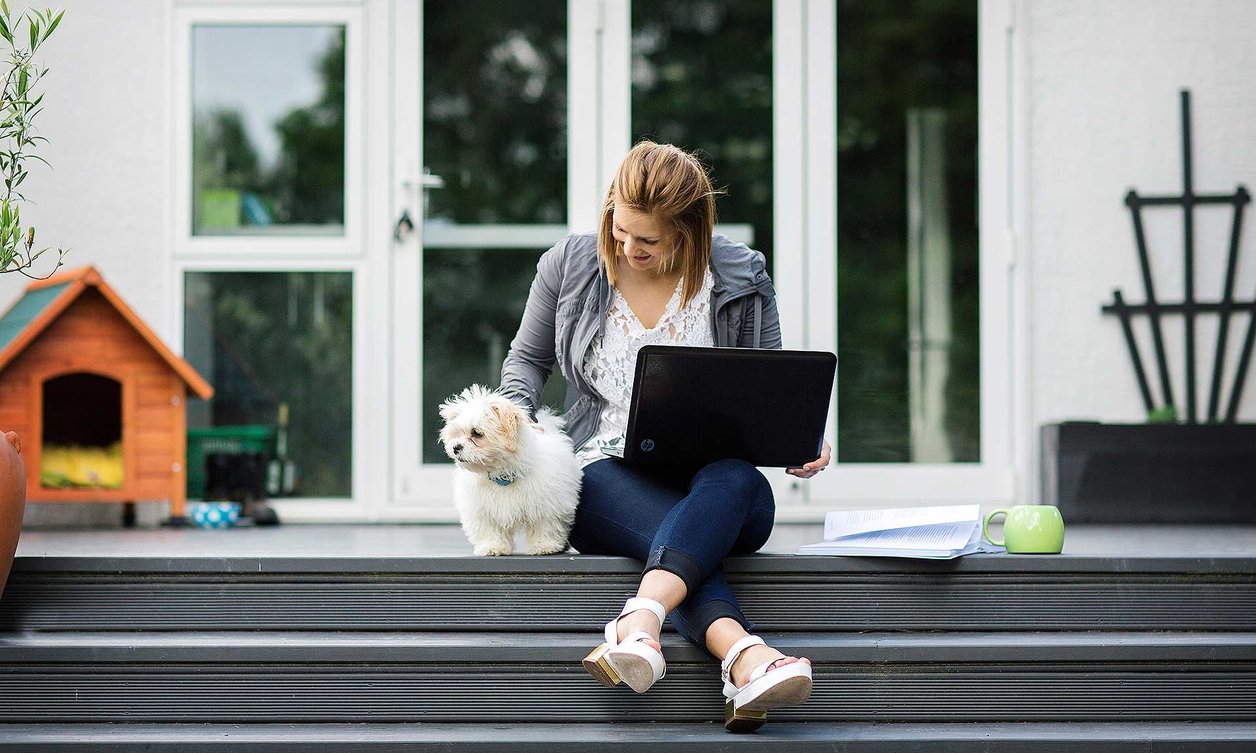 Person sitting on the top step of a deck with a laptop balanced on their knees, patting a small white furry dog